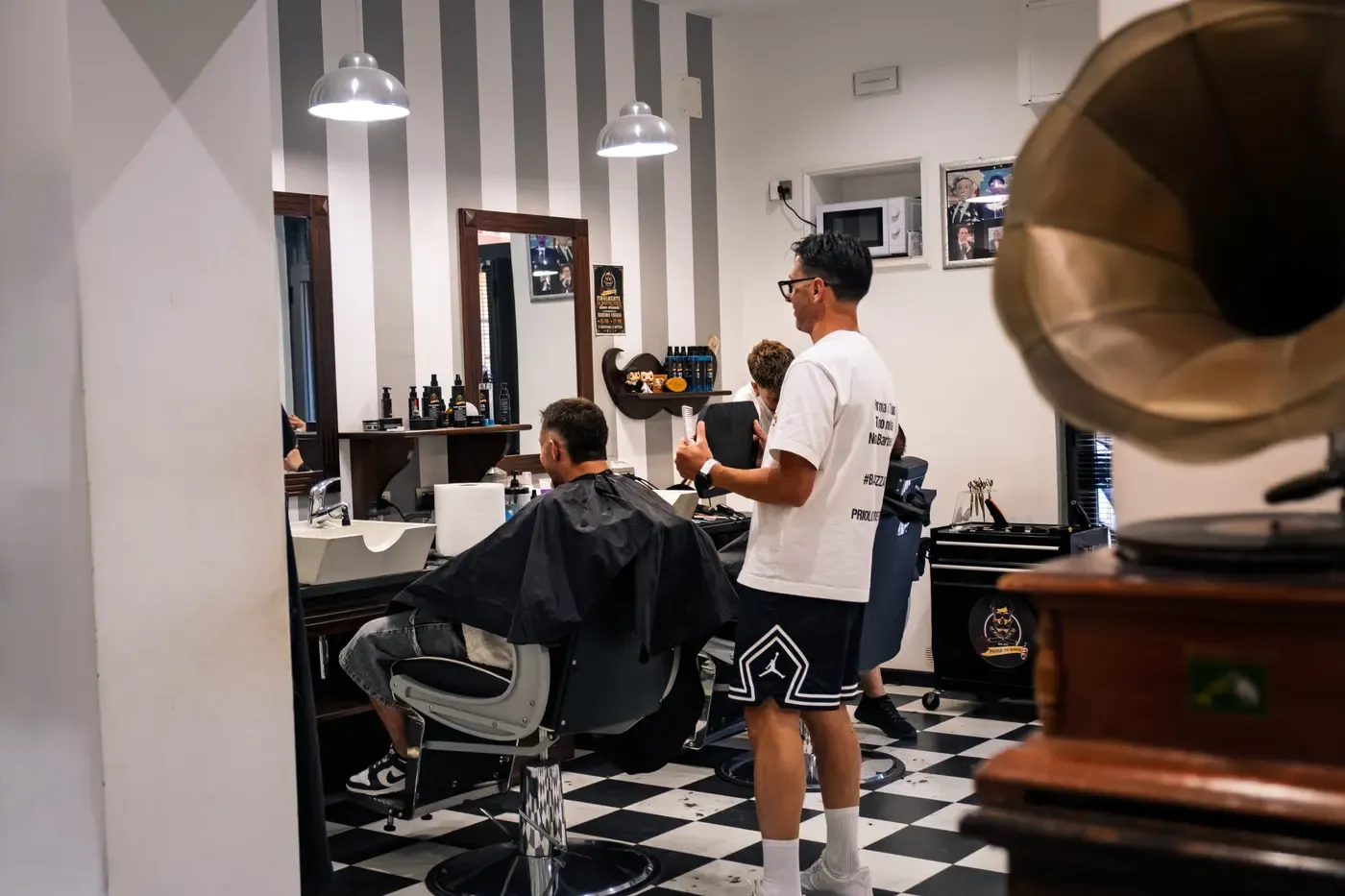 La sala di Priolo the Barbers vista dall'ingresso: pavimento a scacchi, parete a righe, due barbieri al lavoro e il grammofono a tromba in primo piano.