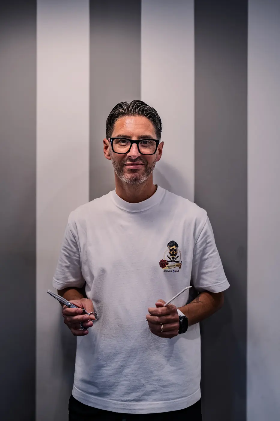 Domenico Priolo, sorridente, con forbici e pettine in mano, davanti alla parete a righe della barberia.