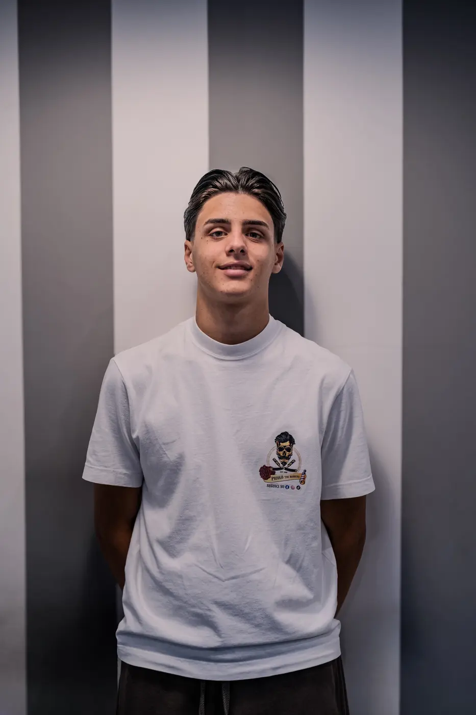 Il figlio di Domenico Priolo, sorridente, davanti alla parete a righe della barberia.